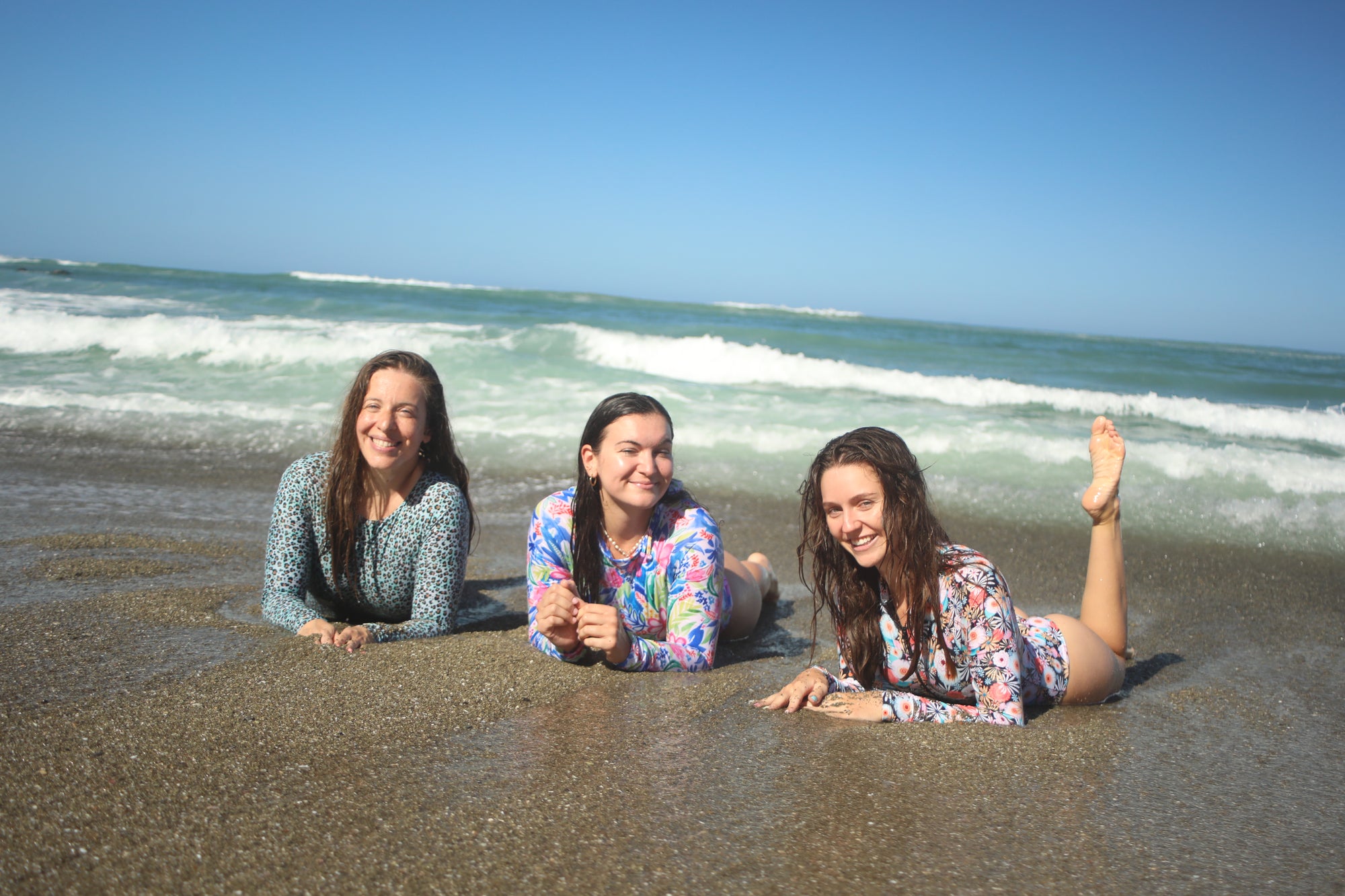 Three women lying on the beach wearing Coconut Gal surf suits with waves in the background